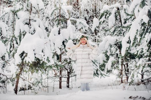 Portrait of a woman in a white fur coat in a cold winter forest. Girl with a wreath on her head in a snow-covered winter forest.