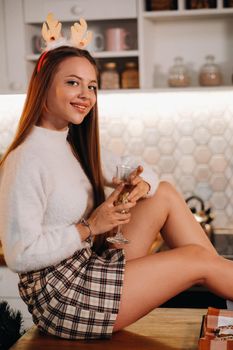 A girl on Christmas day sits on the kitchen table and holds a glass of champagne in her hands.A woman on New year's eve in the kitchen with champagne.