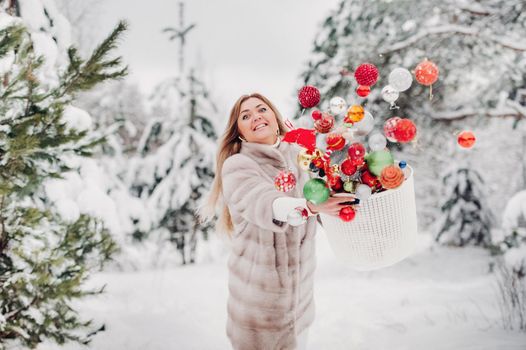 A girl in a fur coat throws Christmas balls to decorate the Christmas tree.Girl throws Christmas decorations from basket into winter forest.