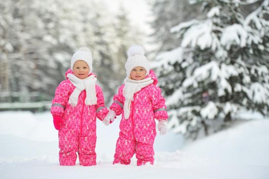 Two little twin girls in red suits stand in a snowy winter forest.