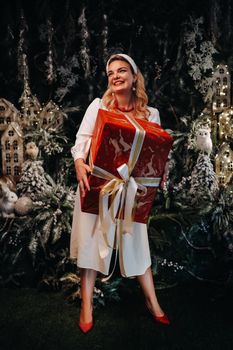 a girl in a white dress with a large Christmas gift in her hands on a fairy-tale background.Smiling woman in white clothes on the background of Christmas trees and small houses.