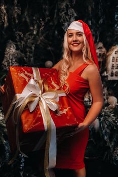 a girl in a Santa hat with a big Christmas gift in her hands on a fabulous Christmas background.A smiling woman in a red dress on the background of Christmas trees and small houses.