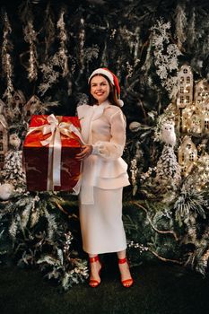 a girl in a Santa hat with a big Christmas gift in her hands on a fairy-tale island.Smiling woman in white clothes on the background of Christmas trees and small houses.