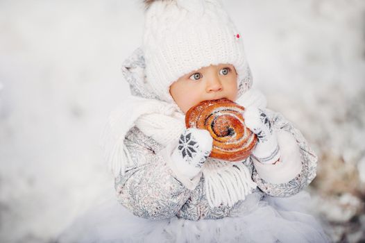 a little girl in white clothes in winter on the street eating a bun.