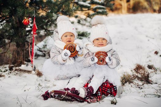 Two little twin girls in white suits sit and eat big buns in the winter outside.