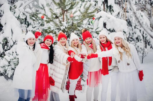 A large group of girls with glasses of champagne in their hands stands in the winter forest.Girls in red and white clothes with new year's drinks in a snow-covered forest