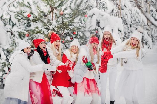 A large group of girls with glasses of champagne in their hands stands in the winter forest.Girls in red and white clothes with new year's drinks in a snow-covered forest