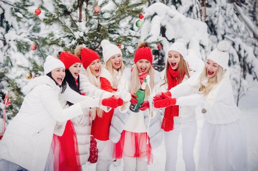 A large group of girls with glasses of champagne in their hands stands in the winter forest.Girls in red and white clothes with new year's drinks in a snow-covered forest