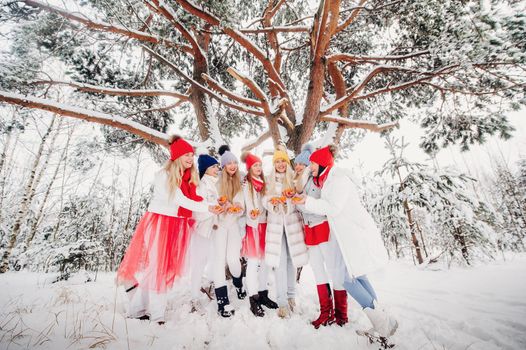A large group of girls with tangerines are standing in the winter forest.Girls in red and white clothes with fruit in a snow-covered forest.