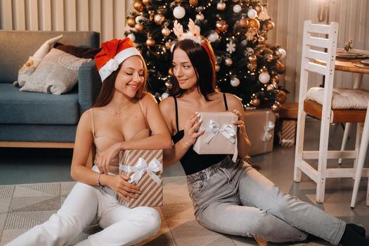 Two beautiful young girls with Christmas gifts in their hands are sitting near the Christmas tree in the house.friends with Christmas dresses and wrapped gifts.