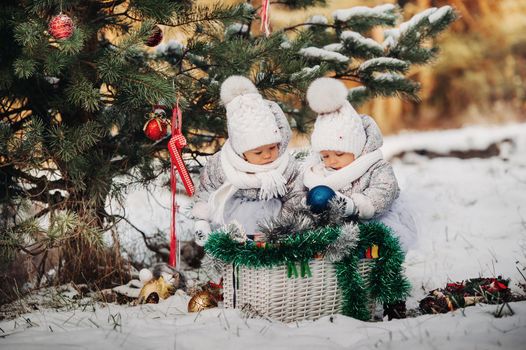 Two little twin girls in white suits take out Christmas balls from a basket near the Christmas tree in winter on the street.