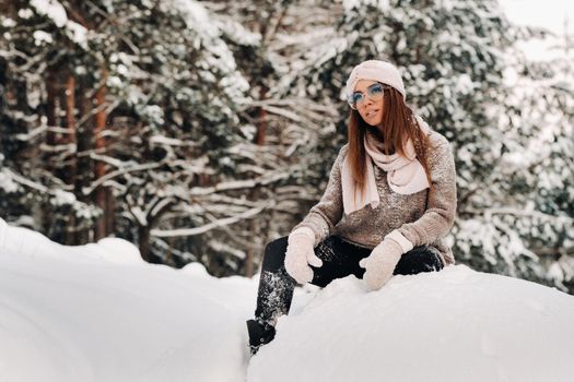 A girl in a sweater and glasses in winter sits on a snow-covered background in the forest.