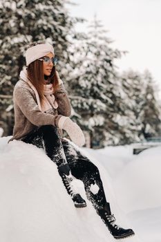 A girl in a sweater and glasses in winter sits on a snow-covered background in the forest.