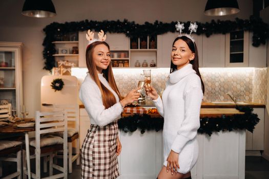 two girls in a cozy home environment with champagne in their hands at Christmas. Smiling girls drink champagne on a festive evening.