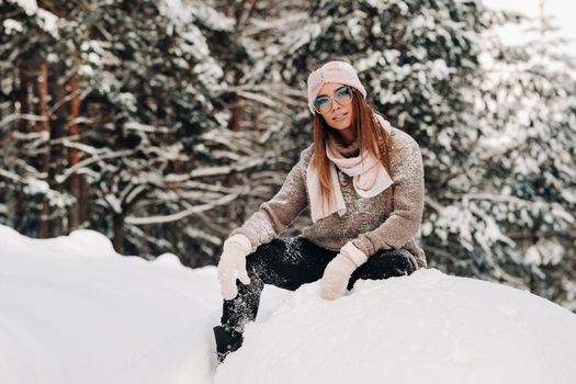 A girl in a sweater and glasses in winter sits on a snow-covered background in the forest.