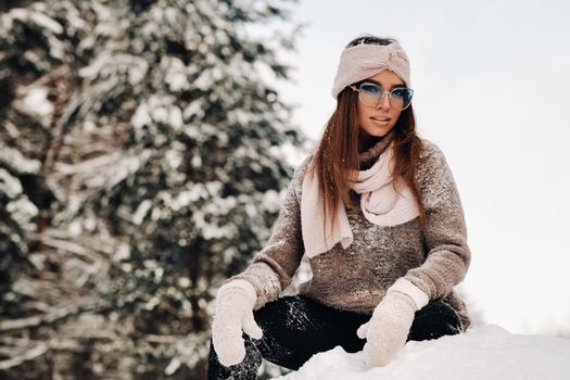 A girl in a sweater and glasses in winter sits on a snow-covered background in the forest.