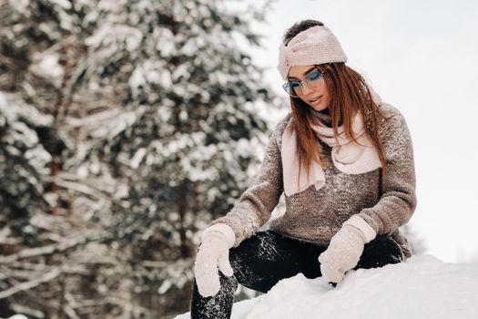 A girl in a sweater and glasses in winter sits on a snow-covered background in the forest.