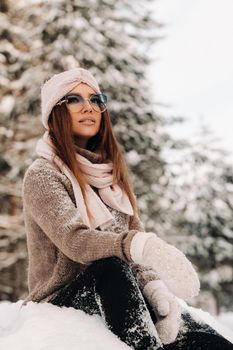 A girl in a sweater and glasses in winter sits on a snow-covered background in the forest.