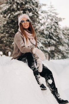 A girl in a sweater and glasses in winter sits on a snow-covered background in the forest.