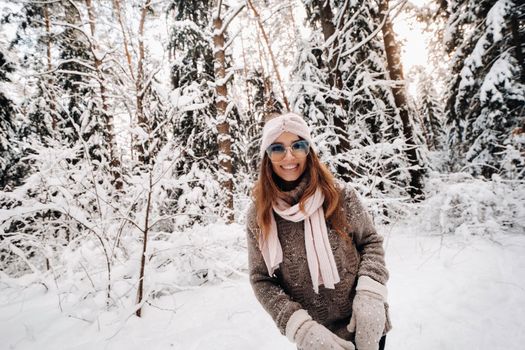 A girl in a sweater and glasses walks in the snow-covered forest in winter.