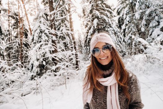 A girl in a sweater and glasses walks in the snow-covered forest in winter.