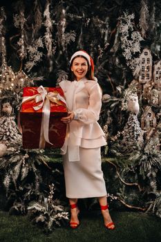 a girl in a Santa hat with a big Christmas gift in her hands on a fairy-tale island.Smiling woman in white clothes on the background of Christmas trees and small houses.