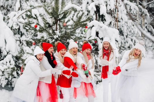 A large group of girls with glasses of champagne in their hands stands in the winter forest.Girls in red and white clothes with new year's drinks in a snow-covered forest