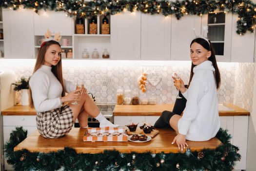 two girls in a cozy home environment in the kitchen with champagne in their hands for Christmas. Smiling girls drink champagne on a festive evening.