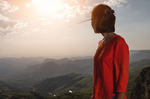 A romantic girl standing on a mountain and admiring the sunrise or sunset over the mountain, the wind develops her hair, a portrait from the side.