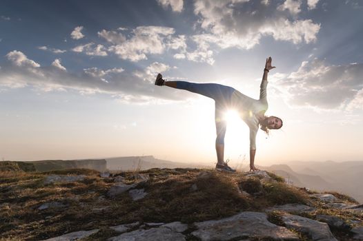 Young woman doing beautiful yoga, sunset or sunrise in mountains over blue sky and clouds against sunlight. Healthy lifestyle successful fitness exercise concept.