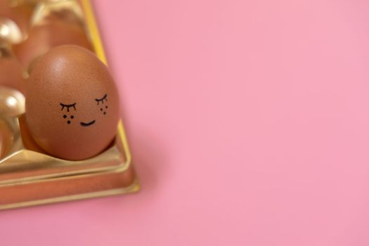 Happy Easter holiday. Cute easter egg in golden box on pastel pink background. Homemade ornament lovely face. Creative arrangement