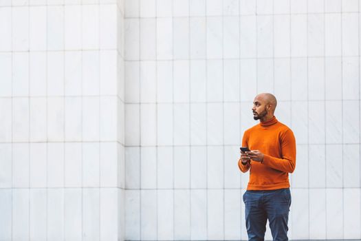 Caucasian man in an orange sweater standing outside in an urban setting holding a digital device, office buildings in the background.