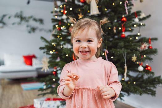 Cheerful young girl, a toddler, enjoying some lollipop while decorating the Christmas tree. Smiling little girl wearing a pink dress and two ponytails.