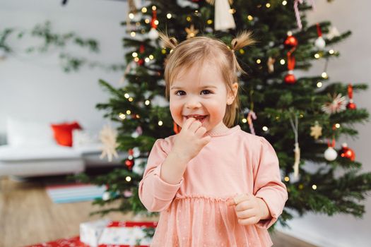 Cheerful young girl, a toddler, enjoying some lollipop while decorating the Christmas tree. Smiling little girl wearing a pink dress and two ponytails.