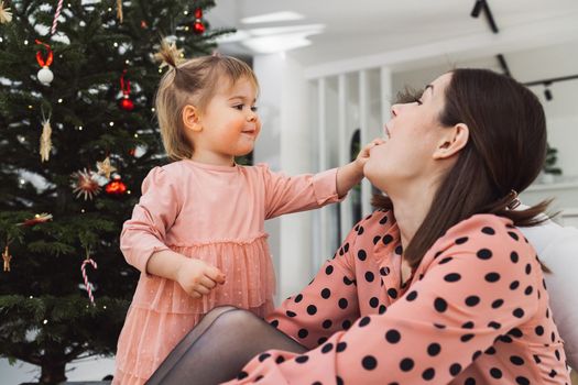 Young caucasian woman, mom, spending time with her toddler, young girl wearing a pink dress matching her mom. Mom and daughter decorating for Christmas together having fun. Mom holding a smiling little girl in her arms.