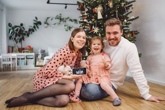 Beautiful young caucasian family Christmas portrait. Mom, dad and a little girl. Mom and daughter wearing pink dresses, dad wearing a white sweater. A little dog, chihuahua joining them on the photos. Family portrait in front of the Christmas tree.