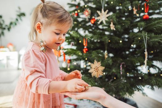 Cheerful young girl, a toddler, enjoying some lollipop while decorating the Christmas tree. Smiling little girl wearing a pink dress and two ponytails.