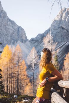 Side view of young caucasian woman hiker in colorful clothes, hiking gear, leaning on a wooden fence somewhere up in the mountains, enjoying the view of the mountain peaks on a beautiful sunny autumn day.