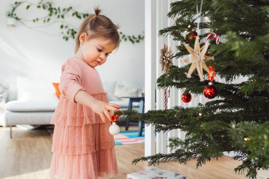 Cheerful young girl, a toddler, enjoying some lollipop while decorating the Christmas tree. Smiling little girl wearing a pink dress and two ponytails.