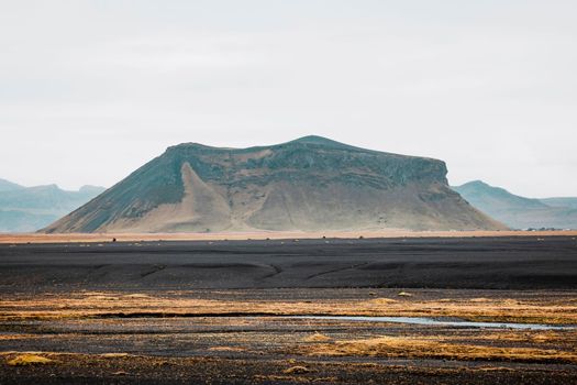 Black volcanic sand beaches in Iceland, a view towards the mainland with a river running trough and some yellow foliage around the water. A mountain right in front.