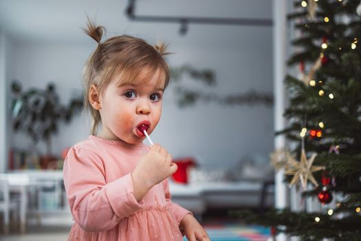 Cheerful young girl, a toddler, enjoying some lollipop while decorating the Christmas tree. Smiling little girl wearing a pink dress and two ponytails.