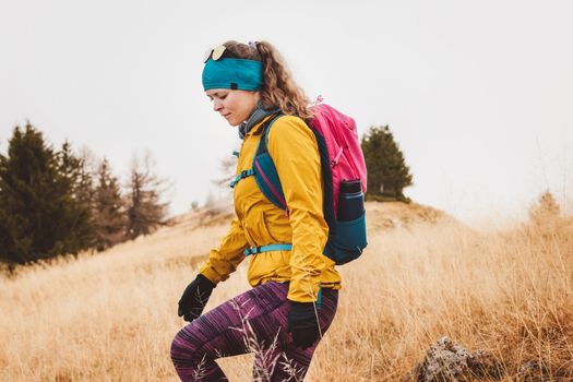 Caucasian woman hiker in yellow jacket and pink backpack hiking in the mountains on an autumn day, wearing gloves and headband.