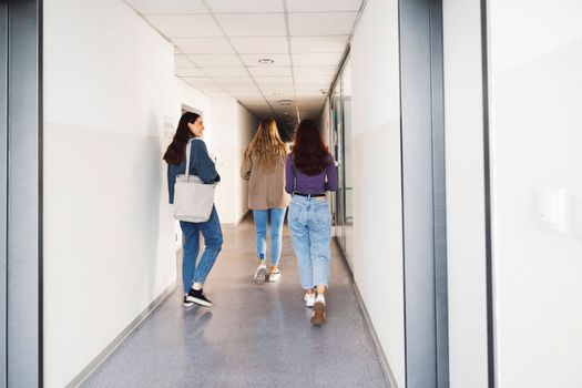 Group of three friends, young caucasian women students walking inside the school during a break, headed to another classroom. Students carrying their books with them.
