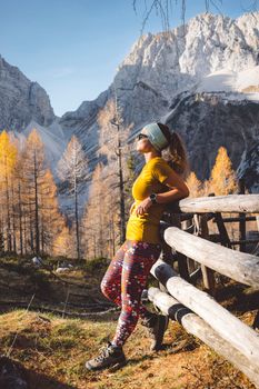 Side view of young caucasian woman hiker in colorful clothes, hiking gear, leaning on a wooden fence somewhere up in the mountains, enjoying the view of the mountain peaks on a beautiful sunny autumn day.