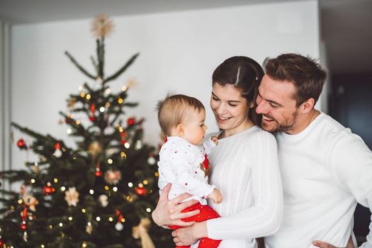 Happy young cheerful caucasian family of three mom dad and baby girl having fun decorating the Christmas tree. Family Christmas portrait.
