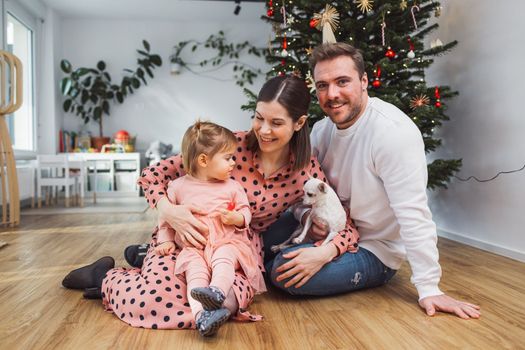 Beautiful young caucasian family Christmas portrait. Mom, dad and a little girl. Mom and daughter wearing pink dresses, dad wearing a white sweater. A little dog, chihuahua joining them on the photos. Family portrait in front of the Christmas tree.