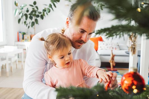 Portrait of young caucasian man with his daughter, decorating the Christmas tree. Loving dad having fun with his little girl on Christmas.