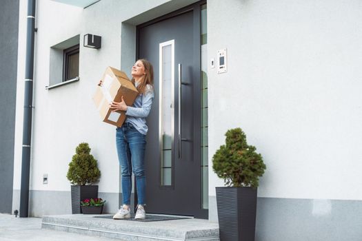 Thrilled caucasian woman standing at the front door with a big cardboard box that just came in the mail. Woman receiving an exciting package, holding it in her arms.