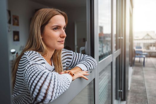 Young blonde caucasian woman looking out the window of her dorm room getting some fresh air on a cold autumn day.