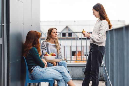 Young caucasian woman college students, sitting on the rood top of their dorm room, eating a snack taking a break from studying, getting some fresh air, on a cloudy autumn day.